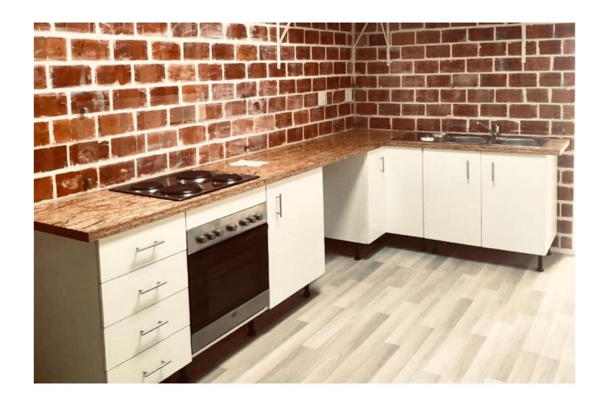 A functional kitchen featuring white base cabinetry, a built-in oven with electric hob, stainless steel sink, and an exposed brick feature wall.