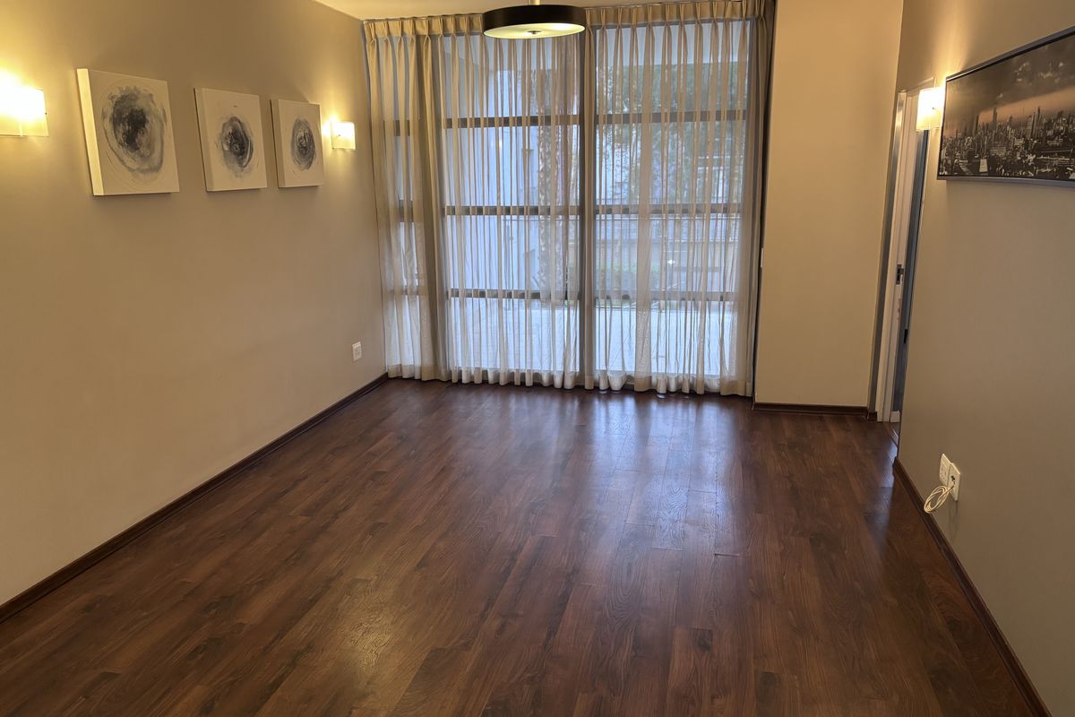 A bright, empty living room featuring dark laminate flooring, floor-to-ceiling windows with sheer curtains, and modern wall-mounted lighting.
