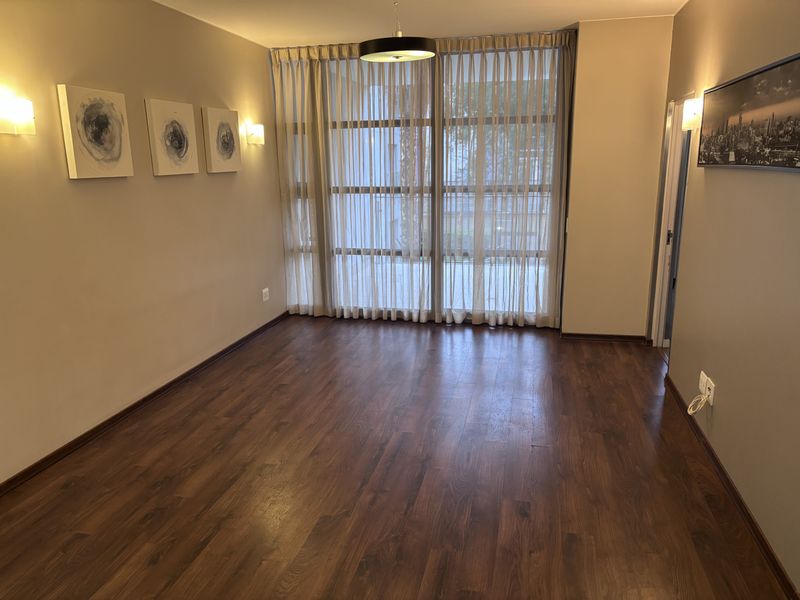 A bright, empty living room featuring dark laminate flooring, floor-to-ceiling windows with sheer curtains, and modern wall-mounted lighting.