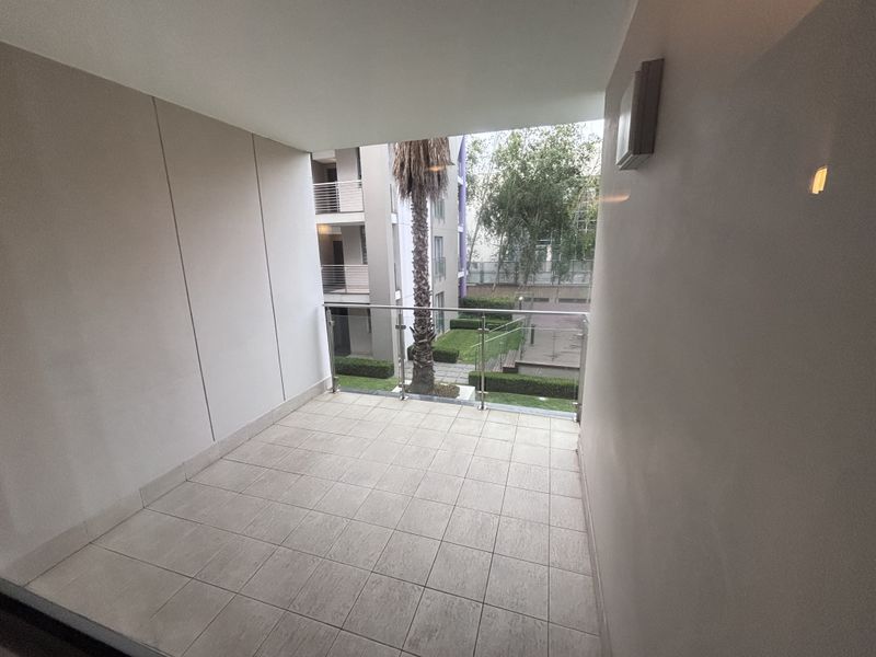 A clean, covered balcony featuring neutral floor tiles and a modern glass balustrade, offering a pleasant view of the complex garden area.