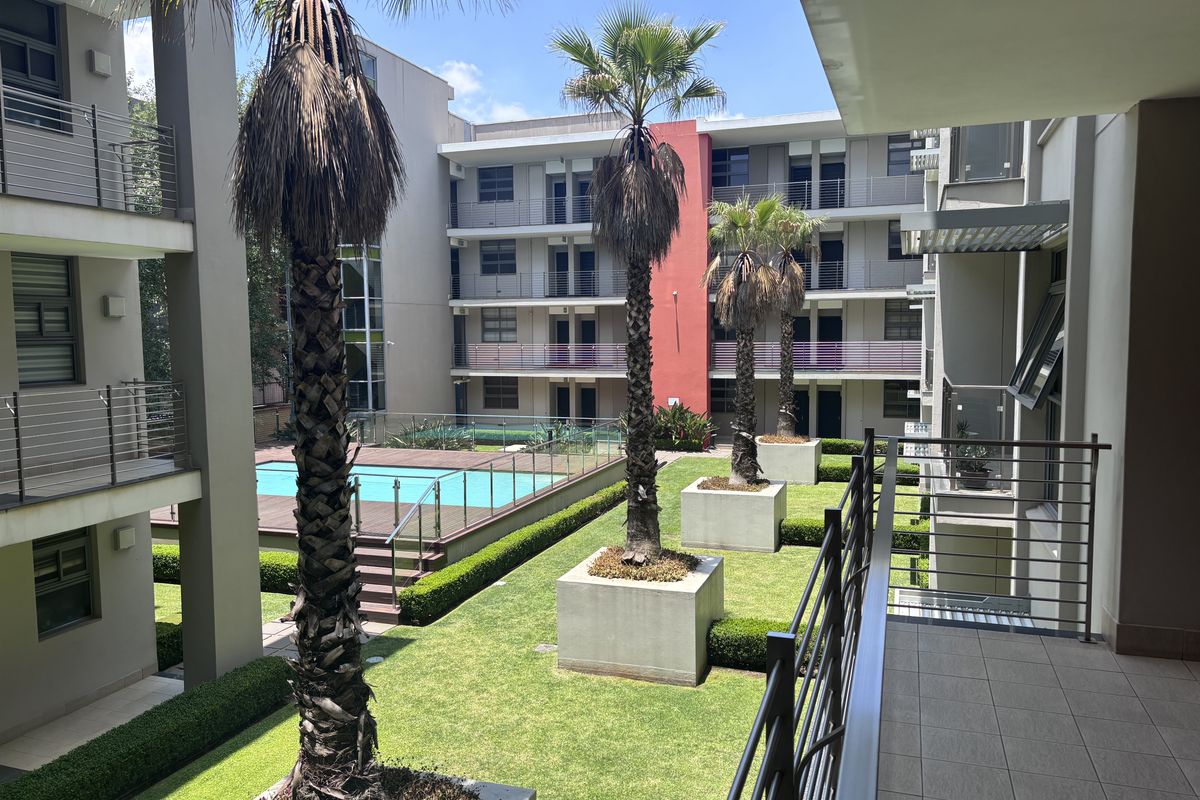 A view from a balcony overlooking a well-maintained apartment complex courtyard featuring a swimming pool, landscaped gardens, and palm trees.