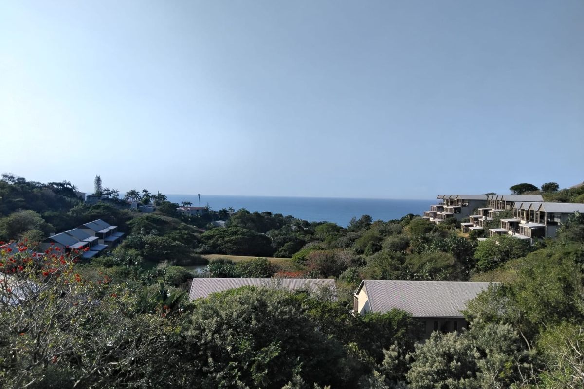 A scenic elevated view overlooking a lush coastal landscape with residential apartment buildings nestled among the trees and the ocean in the distance.