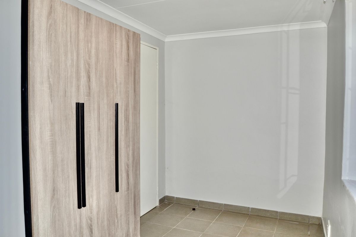 A clean, neutral bedroom featuring built-in wood-grain cabinetry with modern black handles and tiled flooring.