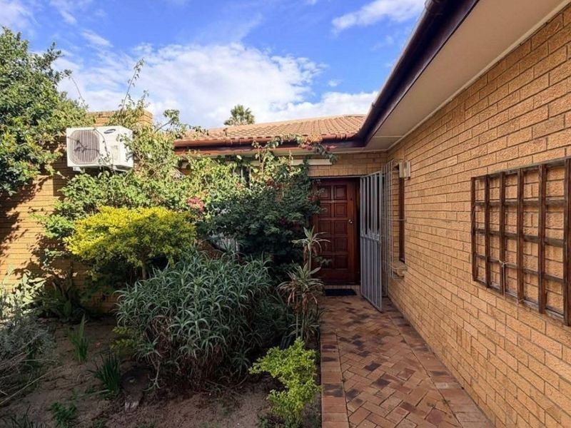 A rear exterior view of the house featuring a brick facade, tiled roof, paved walkway, and established garden area with a security gate.