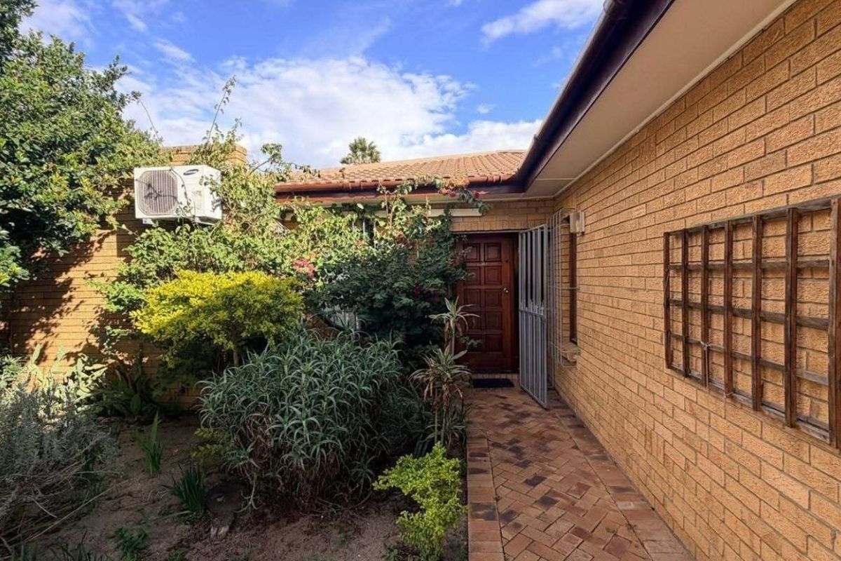 A rear exterior view of the house featuring a brick facade, tiled roof, paved walkway, and established garden area with a security gate.