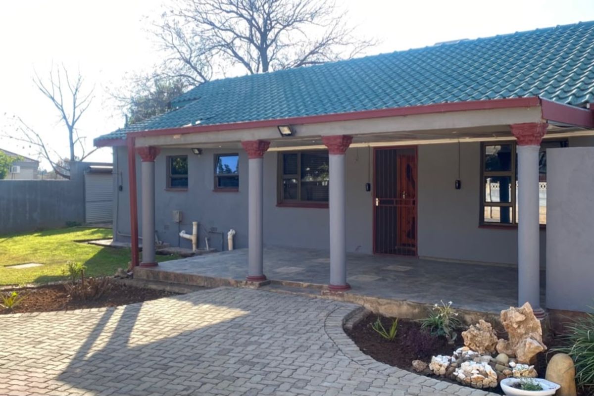A well-maintained rear exterior featuring a covered patio with decorative columns, a tiled roof, and a paved driveway leading to the entrance.