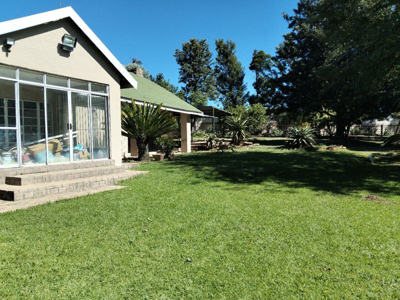 A spacious rear garden featuring a well-maintained lawn, mature trees, and a covered patio area attached to the main house structure.