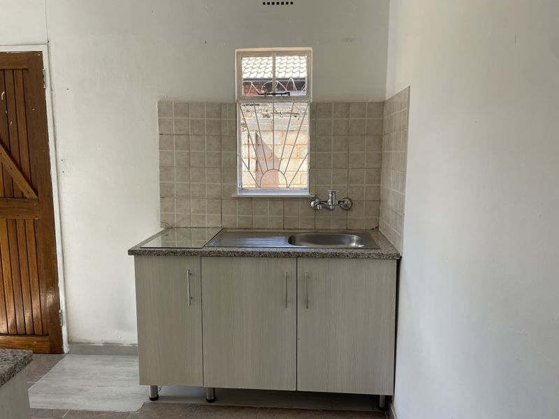 A compact kitchen area featuring a stainless steel sink, laminate countertop, and tiled splashback, set against a plain wall with a security-barred window.
