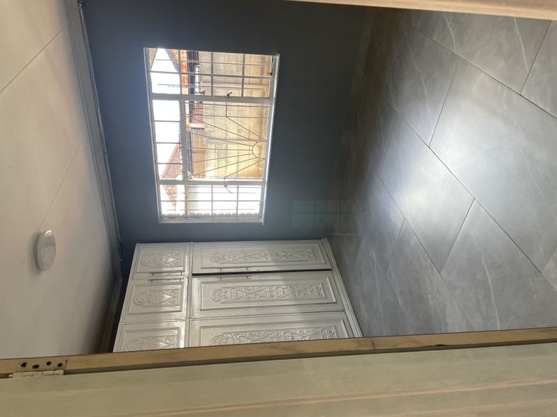 A clean, empty bedroom featuring modern grey floor tiles, built-in white cupboards, and a window fitted with security bars.