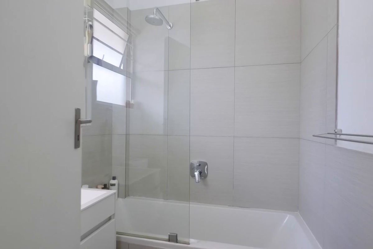 A modern bathroom featuring a bathtub with a glass shower screen, wall-mounted shower head, and neutral tiled walls.
