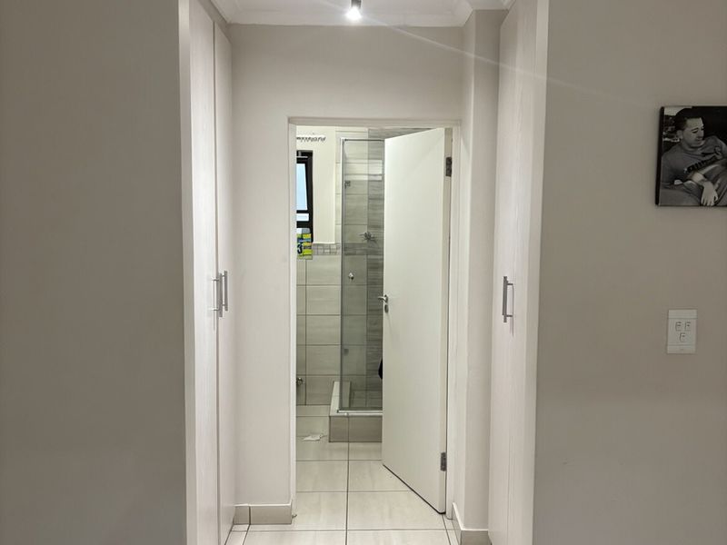 A clean, neutral hallway featuring built-in cupboards and tiled flooring, leading directly into a bathroom with a glass-enclosed shower.