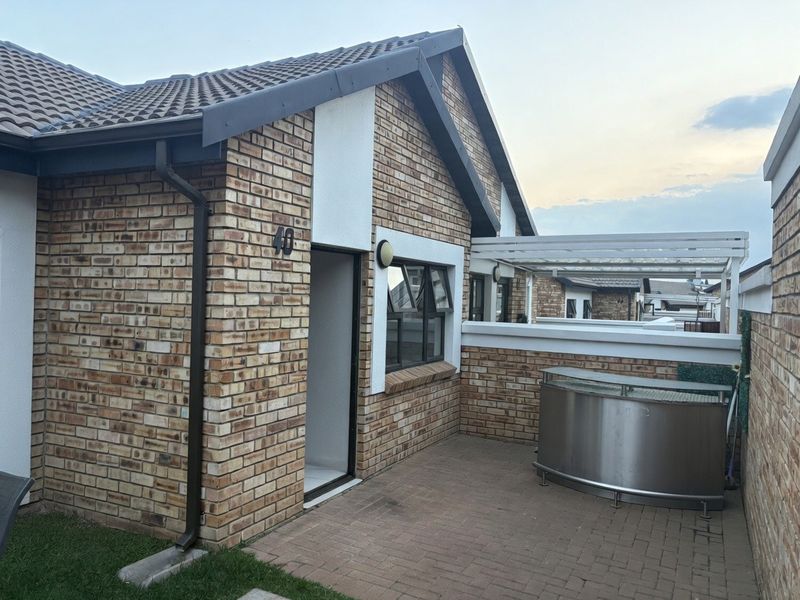 A well-maintained townhouse exterior featuring a face brick facade, tiled roof, paved patio area, and a pergola structure.