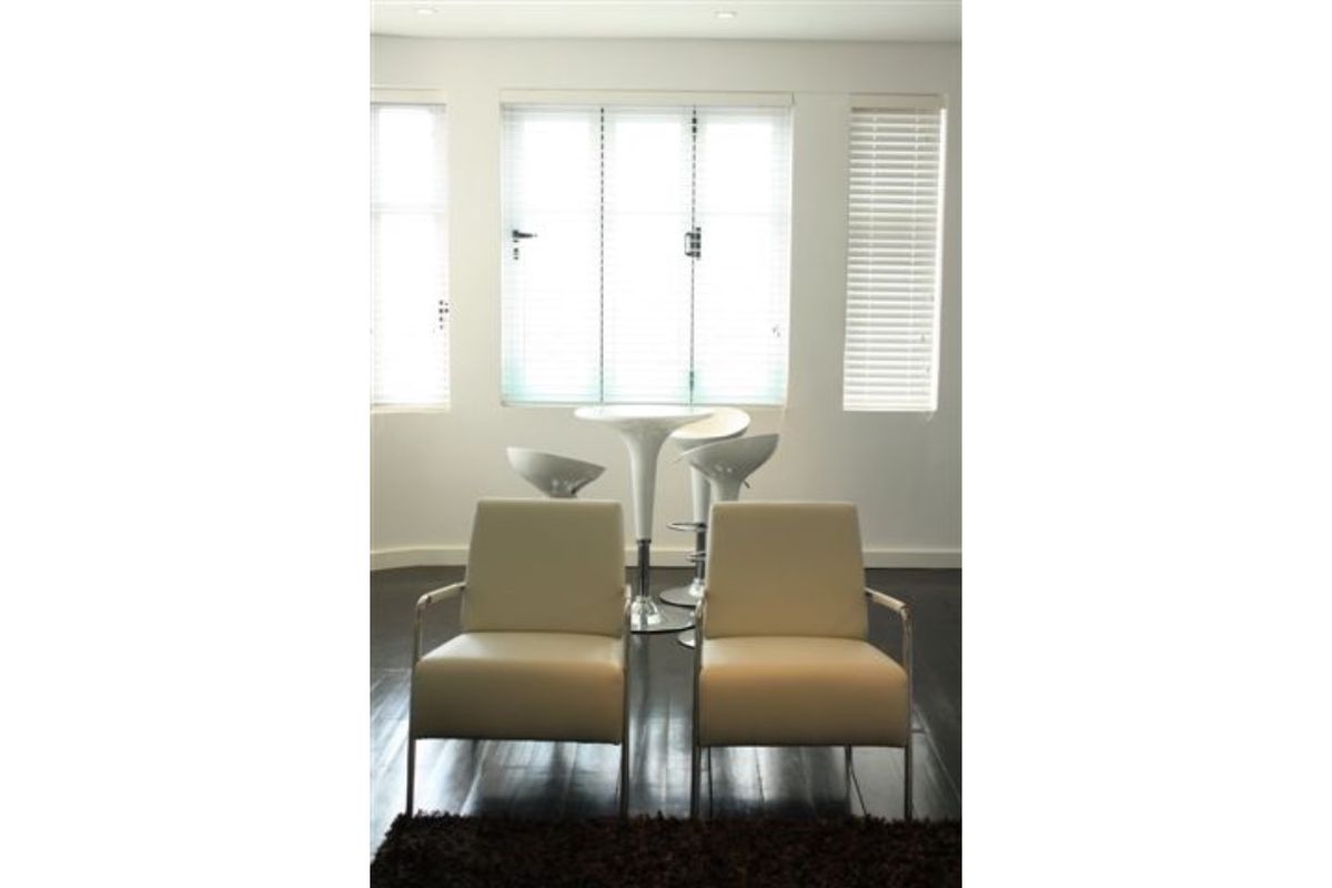 A bright living area featuring dark timber flooring, recessed downlighting, and large windows fitted with white venetian blinds.