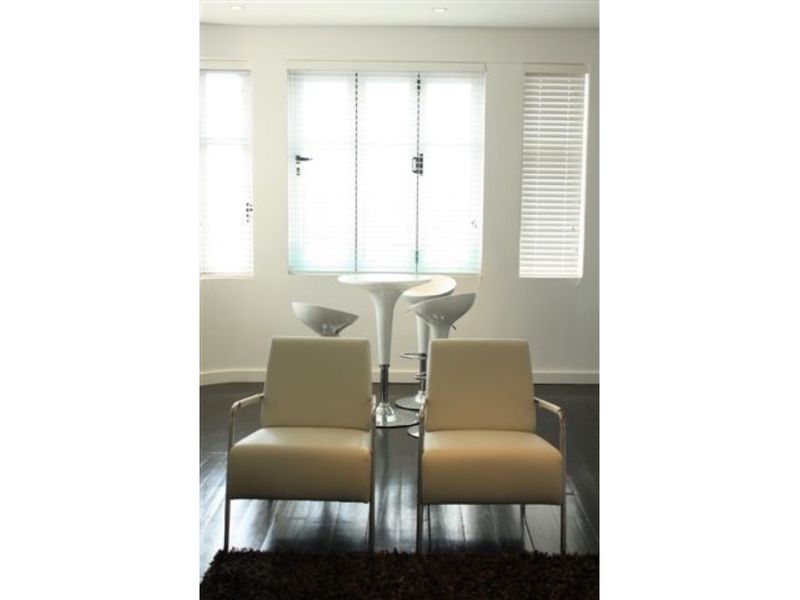 A bright living area featuring dark timber flooring, recessed downlighting, and large windows fitted with white venetian blinds.
