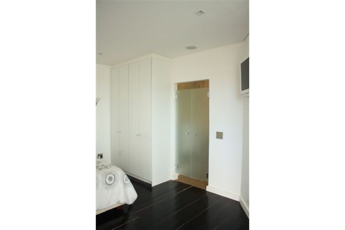 A modern bedroom featuring dark timber flooring, floor-to-ceiling built-in cupboards, and a frosted glass door leading to an en-suite bathroom.