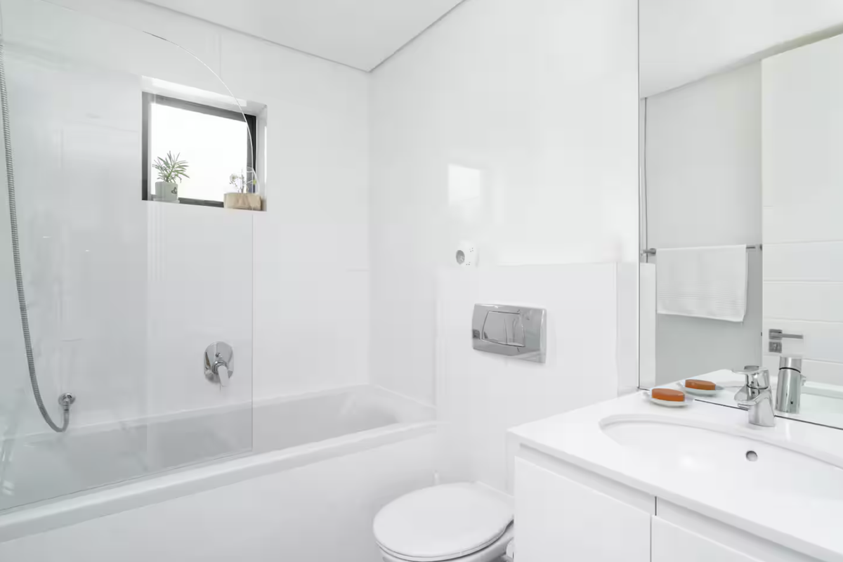 A clean, modern bathroom featuring a white vanity with integrated basin, a bathtub with a glass shower screen, and a wall-mounted toilet.