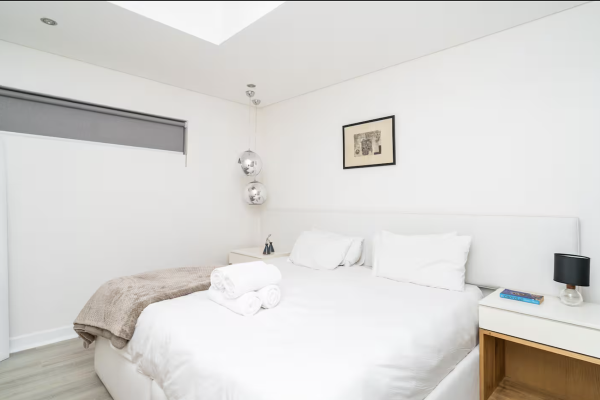 A bright and modern bedroom featuring a built-in headboard, recessed ceiling lighting, and a large window with a fitted roller blind.
