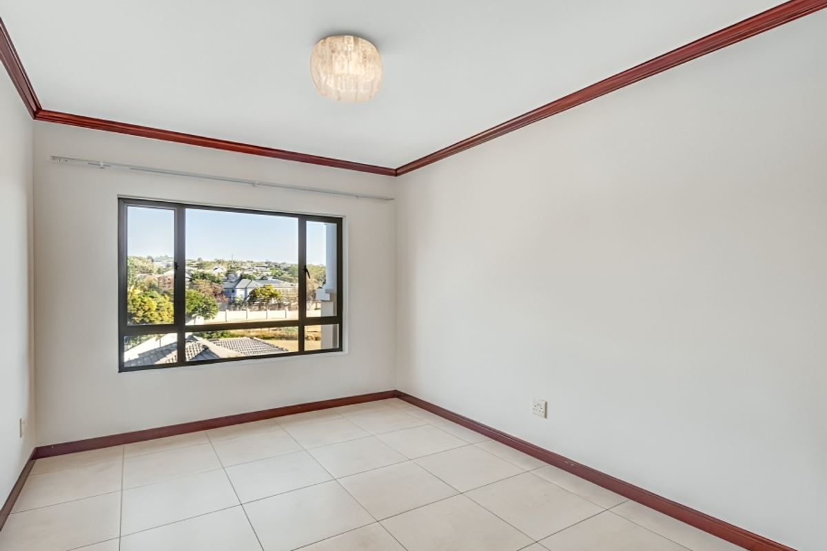 An empty bedroom featuring tiled flooring, dark wood-tone skirting and cornice, and a large window offering an elevated neighbourhood view.