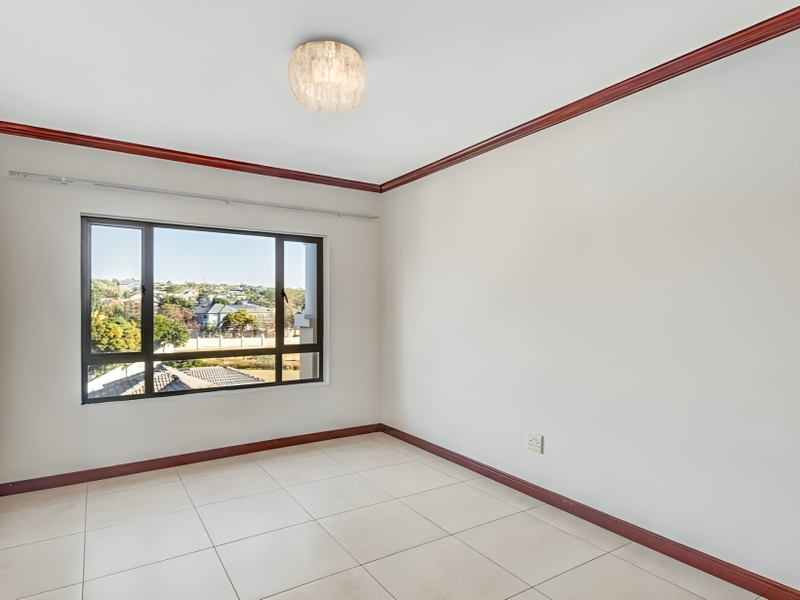 An empty bedroom featuring tiled flooring, dark wood-tone skirting and cornice, and a large window offering an elevated neighbourhood view.