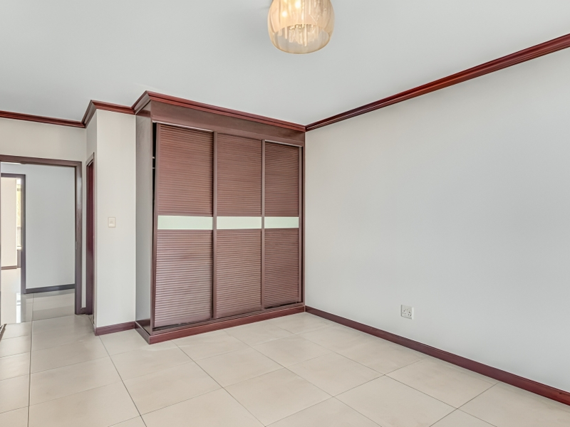 A bright, empty bedroom featuring neutral tiled flooring, dark wood crown molding, and a large built-in wardrobe with sliding doors.