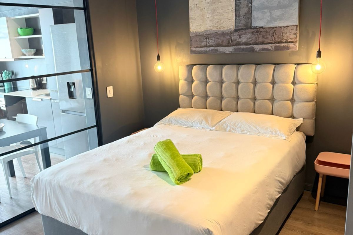 Modern bedroom featuring a concrete ceiling, dark accent wall, and a sleek black-framed glass partition separating the space from the kitchen area.
