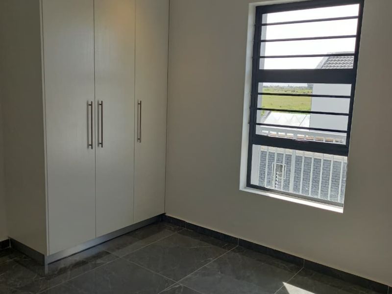 A bright, modern bedroom featuring sleek built-in cupboards, polished grey marble-look floor tiles, and a window fitted with security bars.