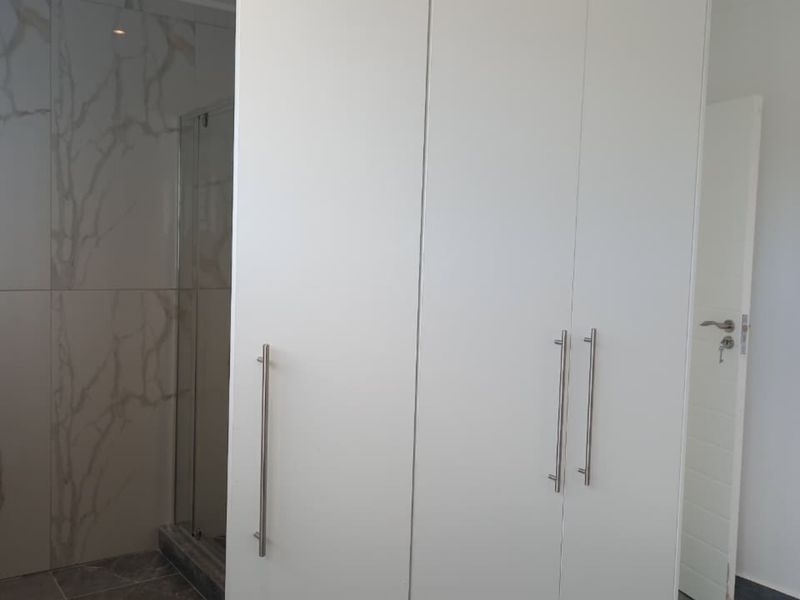 A modern bedroom featuring a large built-in wardrobe and an en-suite bathroom area with marble-look wall tiles and a glass shower enclosure.