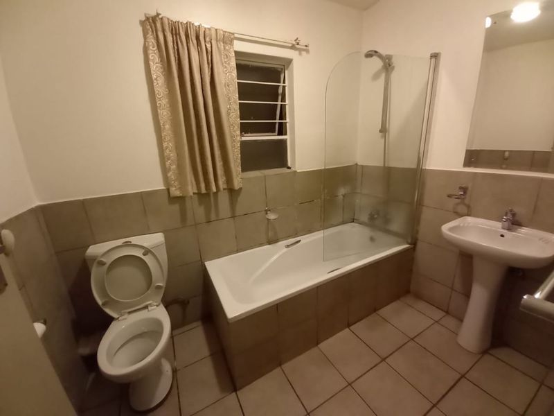A functional bathroom featuring a pedestal basin, toilet, and a bathtub with a glass shower screen, finished with neutral wall and floor tiling.