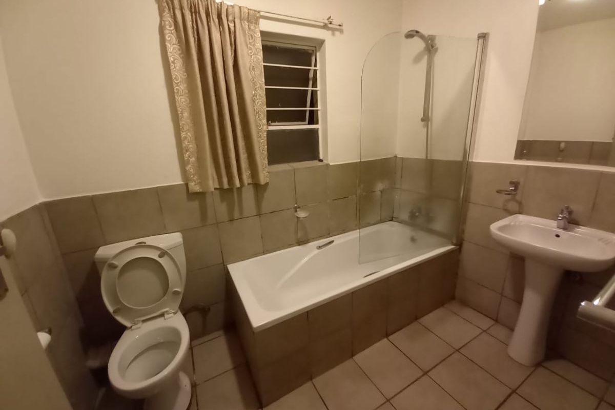 A functional bathroom featuring a pedestal basin, toilet, and a bathtub with a glass shower screen, finished with neutral wall and floor tiling.