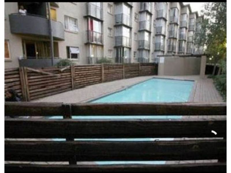 Communal swimming pool area featuring a paved deck and timber perimeter fencing, situated within a multi-story apartment complex.