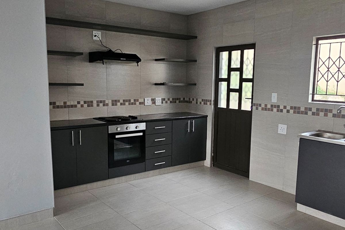 A modern, empty kitchen featuring dark cabinetry, built-in oven and hob, tiled flooring, and neutral wall tiles with a decorative accent border.