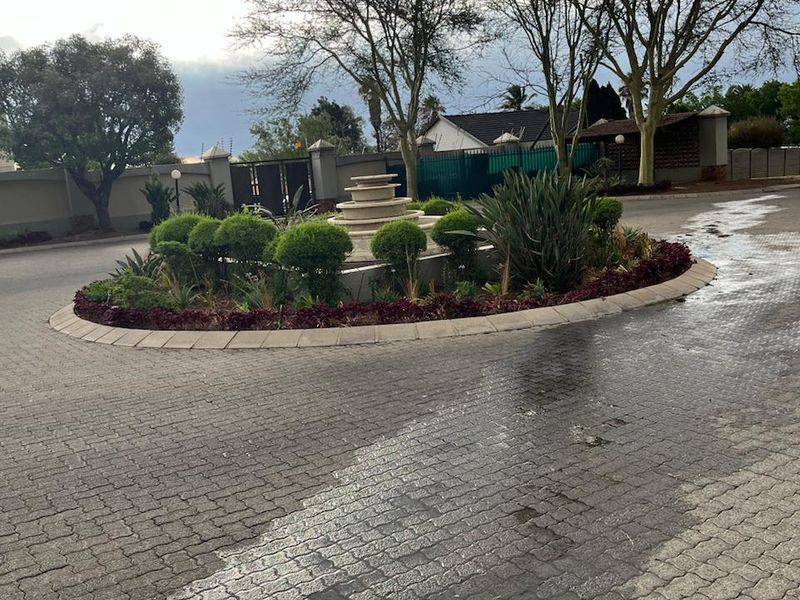 A well-maintained paved driveway entrance featuring a central landscaped garden island with a stone fountain and perimeter security walling.
