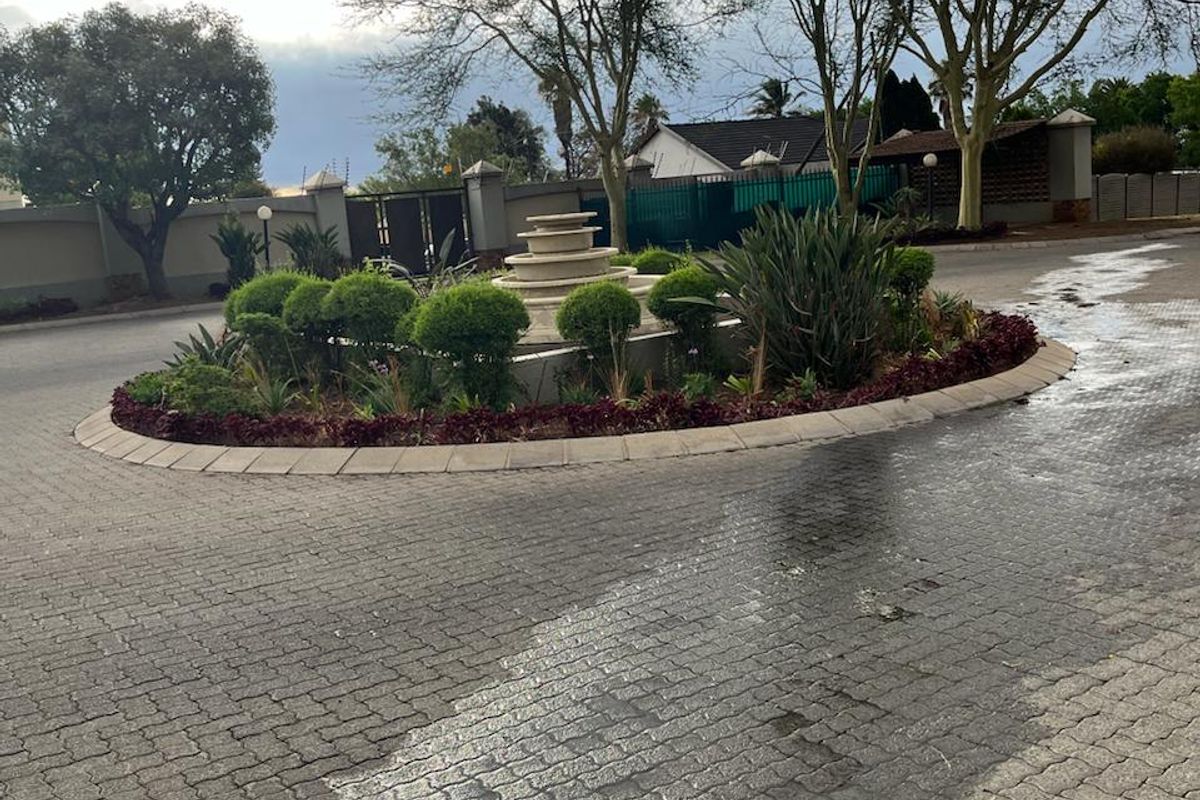 A well-maintained paved driveway entrance featuring a central landscaped garden island with a stone fountain and perimeter security walling.