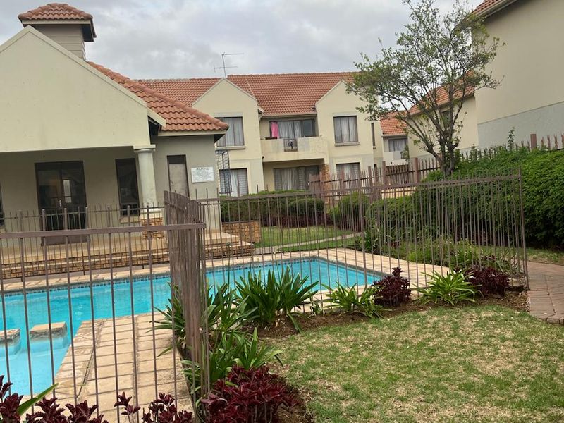 Communal swimming pool area featuring a paved surround, metal fencing, and landscaped garden beds within a residential complex.