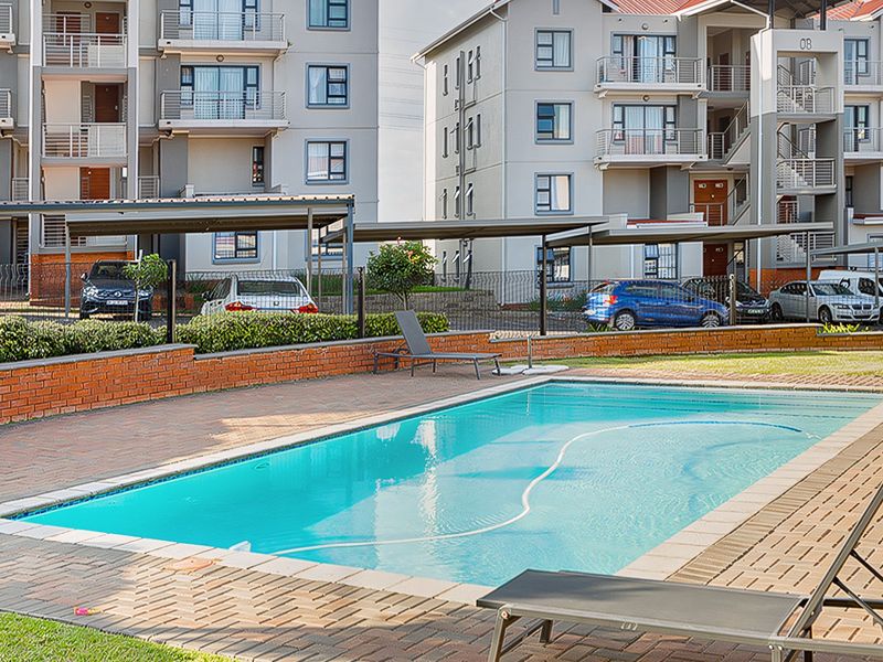 A well-maintained communal swimming pool area featuring brick paving, surrounded by apartment buildings and covered parking bays.