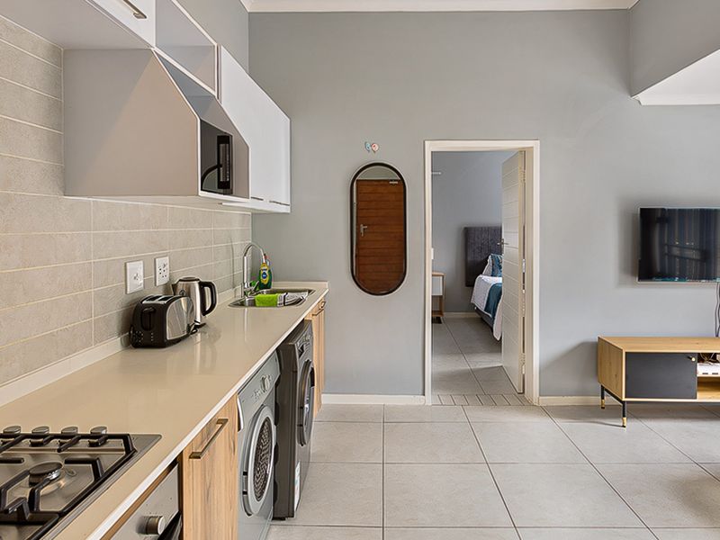 Modern open-plan living area featuring a compact kitchen with gas hob, built-in oven, stone countertops, and tiled flooring, leading into a bedroom.