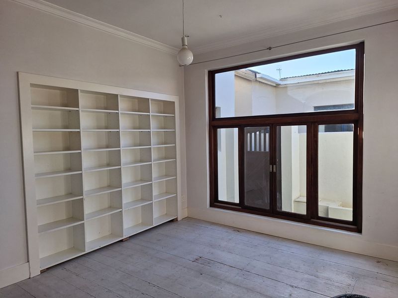 A bright, empty living space featuring built-in white shelving, large timber-framed windows, and original timber flooring with classic cornice detailing.