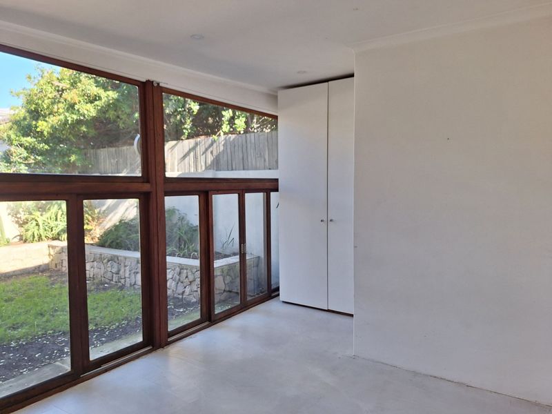 A bright, empty bedroom featuring large timber-framed windows overlooking the garden and convenient built-in cupboards.