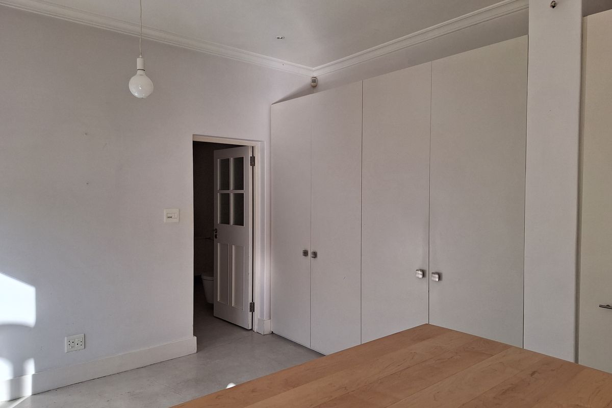 A bright, empty bedroom featuring neutral walls, tiled flooring, and ample built-in cupboard storage with a connecting door to an adjacent room.