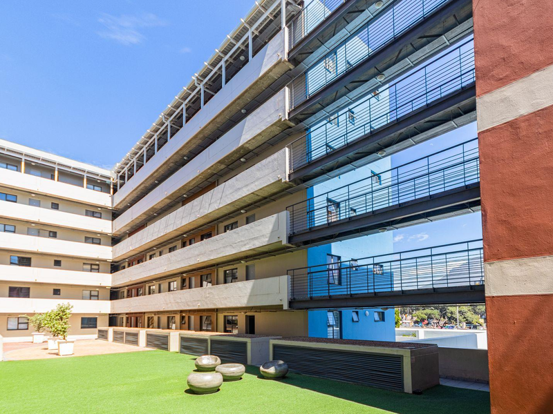 A view of the apartment complex exterior featuring multi-level metal walkways, concrete facade elements, and a landscaped courtyard area with artificial turf.