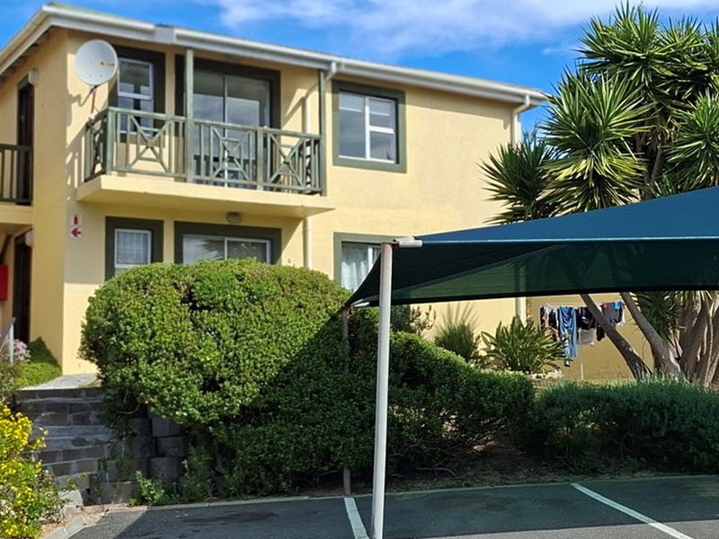Rear view of a two-story apartment building featuring a balcony with railings, sliding doors, and a shaded parking area.