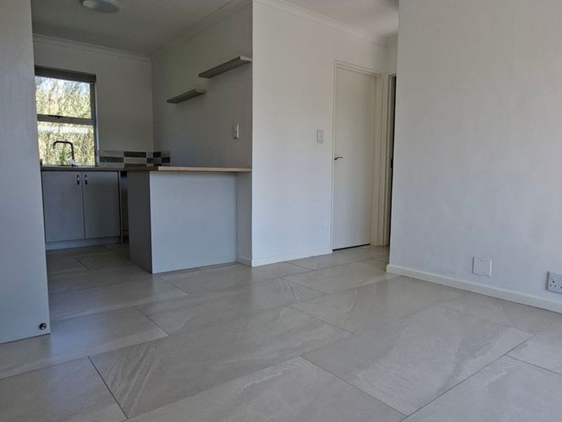 An open-plan living and kitchen area featuring neutral tiled flooring, modern cabinetry, and a breakfast bar in a clean, vacant space.
