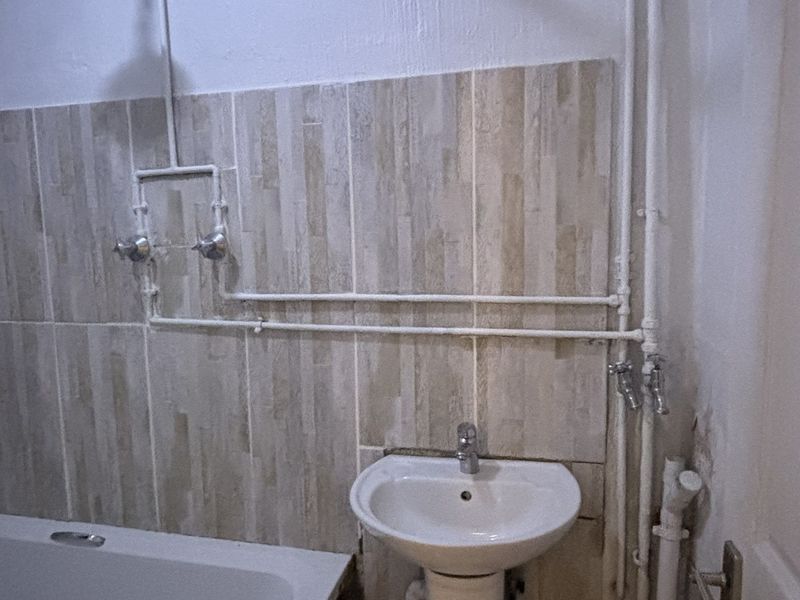 A dated bathroom featuring a pedestal basin, bathtub, and exposed surface-mounted plumbing, requiring cosmetic updates and finish repairs.