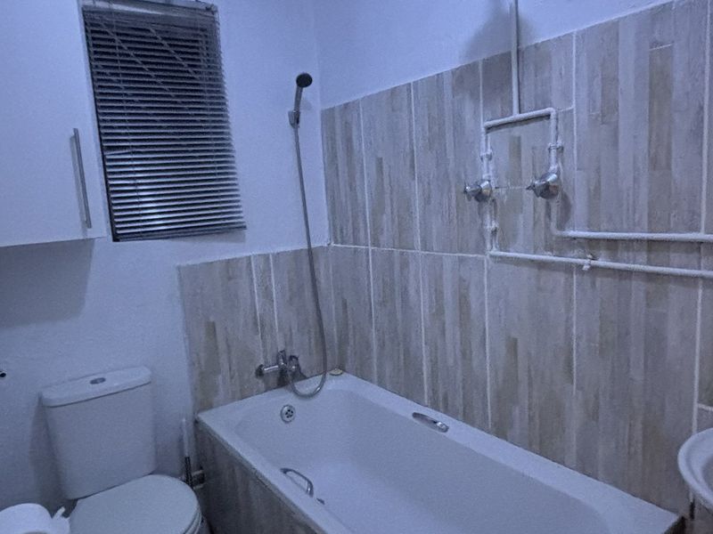 A functional bathroom featuring a bathtub with shower attachment, toilet, and wall-mounted basin, requiring cosmetic updates to finishes and exposed plumbing.