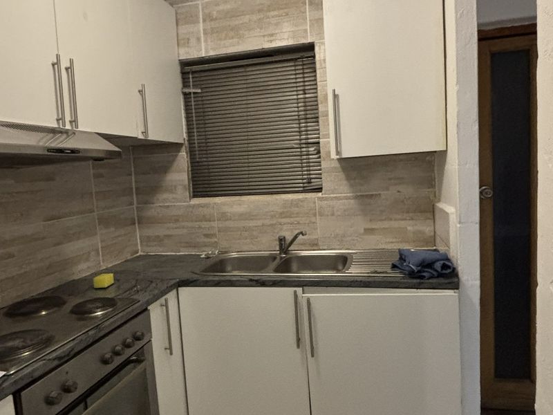 Compact kitchen featuring white cabinetry, stainless steel sink, built-in oven, electric hob, and tiled backsplash with a window for natural light.