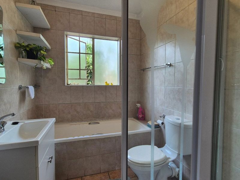 A functional bathroom featuring a built-in bathtub, vanity cabinet, shower enclosure, and neutral ceramic tiling throughout, offering a practical layout with natural light.