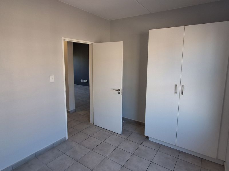 A clean, empty bedroom featuring neutral grey walls, tiled flooring, and a white built-in wardrobe with a view into an adjacent room.