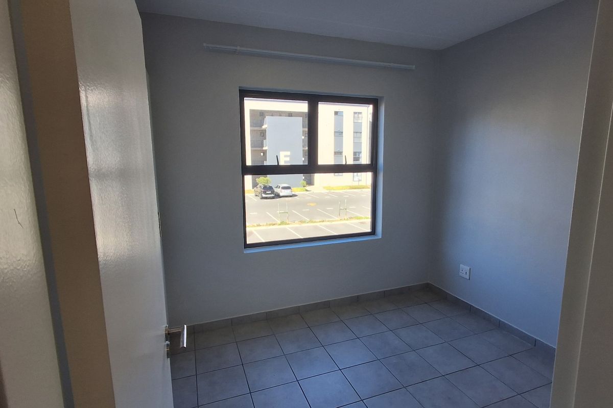 A clean, empty bedroom featuring neutral wall paint, tiled flooring, and a large window providing natural light and views of the complex parking area.