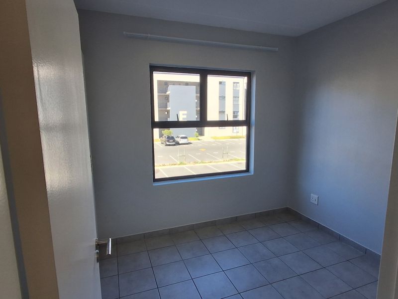 A clean, empty bedroom featuring neutral wall paint, tiled flooring, and a large window providing natural light and views of the complex parking area.