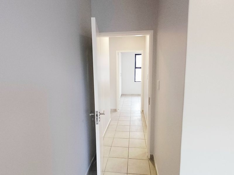 A clean, neutral-toned hallway featuring tiled flooring and connecting internal doorways, leading towards a bright room with a window.